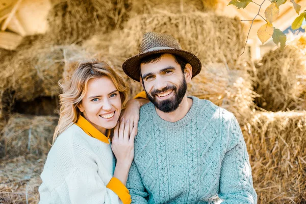 Retrato de una feliz pareja de granjeros mirando la cámara mientras están sentados en pilas de heno en la granja - foto de stock