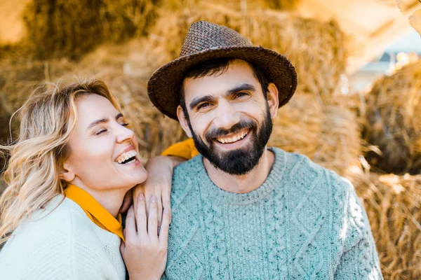 Pareja de agricultores que ríen sentados en pilas de heno en la granja - foto de stock