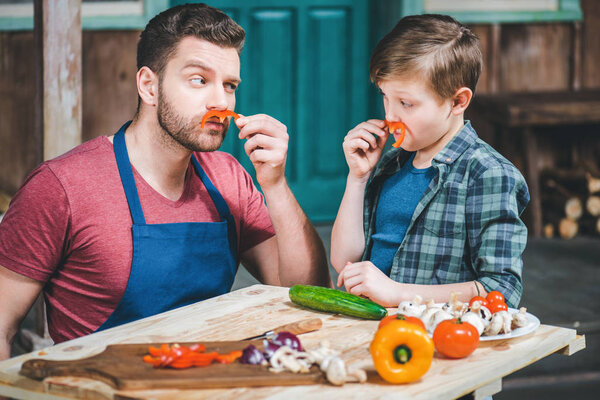 little boy with father doing moustaches with slices of pepper while cooking at table 