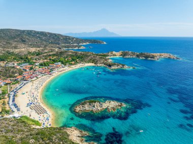 Yunanistan 'ın Sithonia kentindeki Kalamitsi Sahili' nin havadan görünüşü, turkuaz sular, altın kum, sahil şemsiyeleri, bir sahil köyü ve arka planda Athos Dağı bulunuyor.