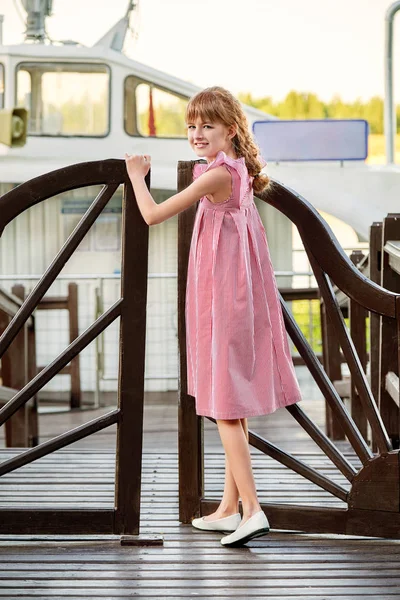 Mladá dáma dítě 11 let stojí poblíž dřevěný most při pohledu kamery, venku. Po celé délce teenager v elegantní letní šaty v červené pruhy pro plavbu, moře, dovolená. Módní kid koncept . — Stock fotografie