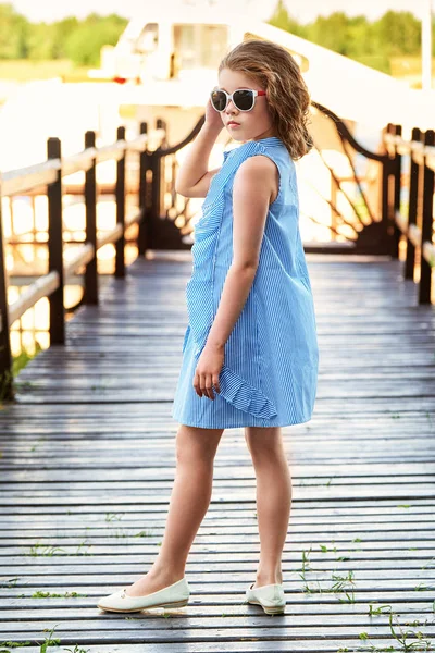 Sevimli küçük kız zarif mavi elbise genç bağlantı noktasında dinlenme için. Güzel çocuk yaz serin giyim, güneş gözlüğü takıyor. Moda çocuk kavramı. Köprü pier ayakta. Çekici genç hanım.