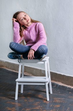 Şık çocuk kız moda poz sandalyede oturuyor. Güzel göz alıcı çocuk teenagerbrunette tanıtır, öylesine, kentsel, sokak, gençlik tarzı. kıyafetler. Stüdyo çekim, çatı katı iç.