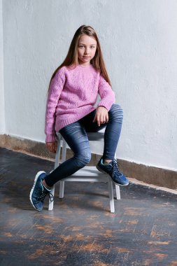 Moda poz, beton beyaz arka plan iskemlede kız kendine güvenen çocuk. Şık model pembe kazaklı. Güzel göz alıcı çocuk genç giyim rahat, kentsel tarzı. Stüdyo vurdu.