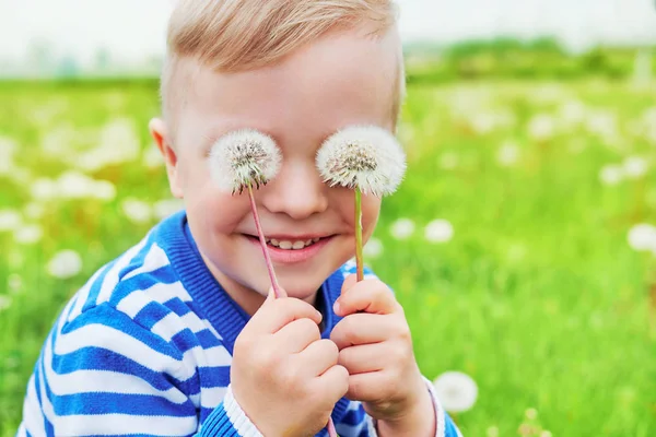 Happy face kid smile. Close up portrait joy child outdoors. Little boy playful smiling holding dandelions on eyes as eyeglasses. Joyful childhood, summer day. Background green grass.