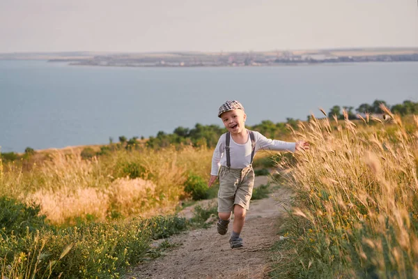 Šťastné dítě chlapec běží na louce v létě v přírodě. Malé dítě hraje jarní den na poli. Krásné usměvavé dítě nosit módní retro oblečení, čepice, šle. — Stock fotografie