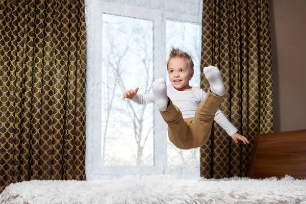 Dětské zábavné domů. Veselý kluk v pohybu, skákání, směje se na posteli. Malé dítě 6 let šťastně hraje ráno v pokoji. — Stock fotografie