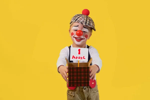Kid clown with red nose 5 years on the bright yellow background. Little child holds box with white sheet, text 1 April. Concept birthday, holiday, celebration, party. Happy jester looking small box.
