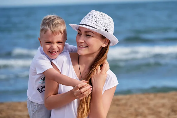 Szczęśliwy synka chłopak przytula matki. Dziecko kocha Mama, ona wygląda odległości i uśmiecha się. Portret młodej kobiety w kapeluszu wraz z cute blond dziecko po odpoczynku na plaży. pojęcie holiday, wakacje. — Zdjęcie stockowe