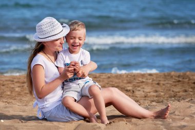 Genç anne oğlu ile sahilde oturuyor. Şirin anne birlikte çocuk çocuk kucaklayan, gülmek gülümseyen, yaz günü. Mutlu çocukluk oynarken açık kum, deniz, neşe, eğlenceli kaygısız. tatil, tatil kavramı.
