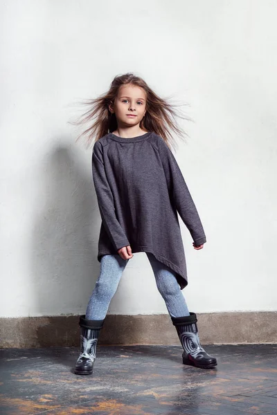 Niño posando en una ropa elegante, urbana y casual. niña de moda de pie cerca de la pared blanca. Retrato joven y encantador niño de moda en leggings, suéter, botas de goma . — Foto de Stock