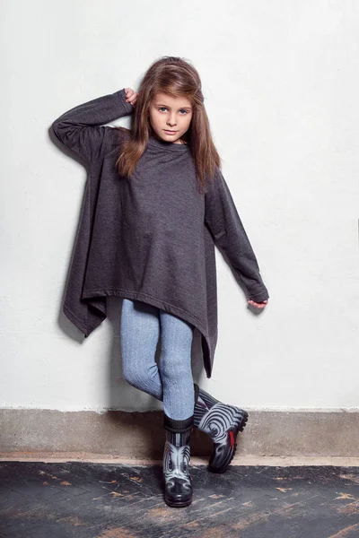Moda model młody dzieciak pozowanie w stylowy, miejski, dorywczo ubrań. Małe dziecko wygląda ręką poważne, dotykając jej włosy, stojący w pobliżu biała ściana. Leginsy dla dzieci, swetry, buty gumowe.. — Zdjęcie stockowe