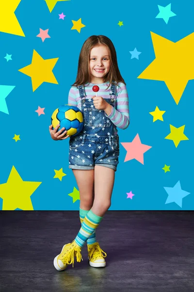 Cute Little Girl w denim przed niebieską ścianę. Koncepcja mody. Dziecko model w dżinsy kombinezon stwarzające w Studio, wielokolorowe gwiazdy tła . — Zdjęcie stockowe