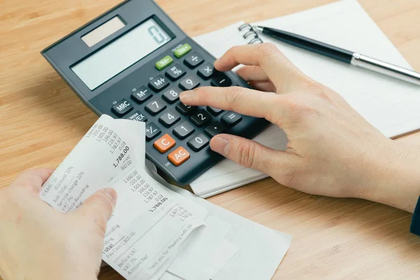 Cost and expense calculation or bill payment concept, hand put finger on calculator and black pen on paper notepad and holding pile of bills or receipt in the left hand on wooden table