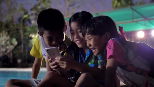 夕方にはスイミングプールの近くでリラックスしたスマートフォンを使用してアジアの子供たちのグループ — ストック動画
