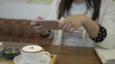 Asyalı kadın kapuçinosunun akıllı telefonuyla fotoğrafını çekiyor..