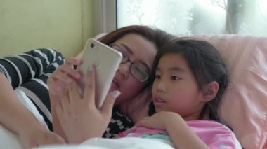 Mutlu Asyalı aile, genç anne ve kızı yatak odasında cep telefonu oynuyorlar komik anne ve güzel çocuklar telefonla eğleniyorlar..