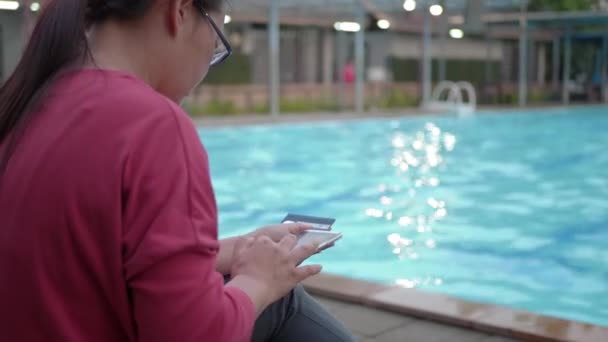 スイミングプールの近くでスマートフォンでオンライン決済を行うアジアの女性 — ストック動画