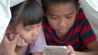 Asyalı çocuklar yataktaki battaniyenin altında akıllı telefon ya da tablet kullanıyorlar..