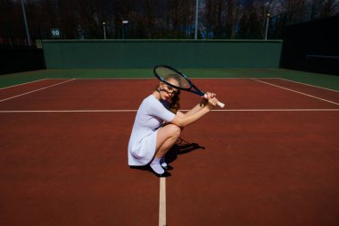 Sahada tenis raketi tutan beyaz elbiseli ve topuklu ayakkabılı seksi kız tenisçi. Genç kadın tenis oynuyor.