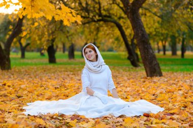 Çiçekli güzel kızıl saçlı bir kızın portresi sonbahar ormanının arka planında beyaz bir elbiseyle çekici görünüyor ve gülümsüyor..