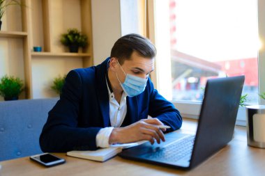 Grip karantinasında yüz maskesi takmış genç yakışıklı iş adamının portresi. Bilgisayarlı, cep telefonlu bir fotoğraf. Koronavirüs salgını.