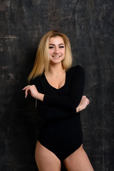 Young plump woman with bright makeup in black bodysuit is posing at dark background, isolated with copy space. Concept of xxxl fashion and junk food