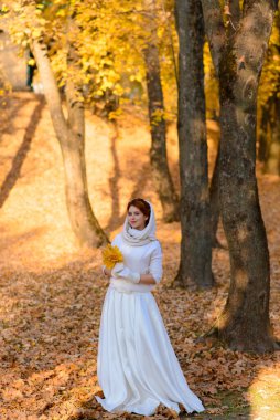 Uzun beyaz gelinlikli şirin kız kırsal yolda poz veriyor altın saatinde ormanda sonbahar ağaçları arasında..