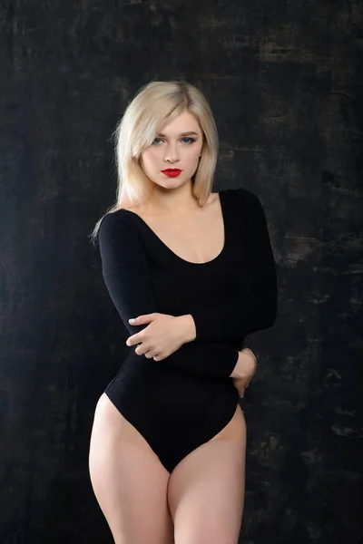 Young plump woman with bright makeup in black bodysuit is posing at dark background, isolated with copy space. Concept of xxxl fashion and junk food