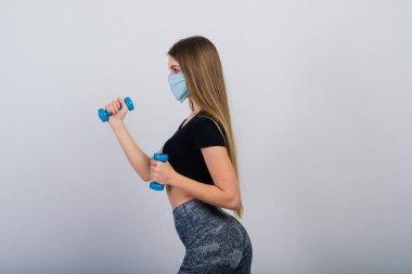 Beyaz arka planda halterleri olan koruyucu maskeli genç bir kadın. Karantinaya alınmış sporlar