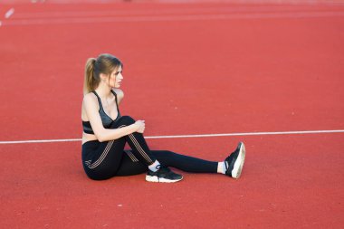 Siyah saçlı bir bayan koç, stadyumun kırmızı koşu pistinde spor üniforması içinde duruyor..