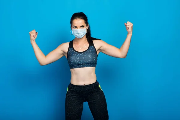 Young strong fitness woman in face mask working out isolated on blue wall background studio.