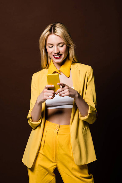 happy stylish blonde girl using yellow smartphone isolated on brown