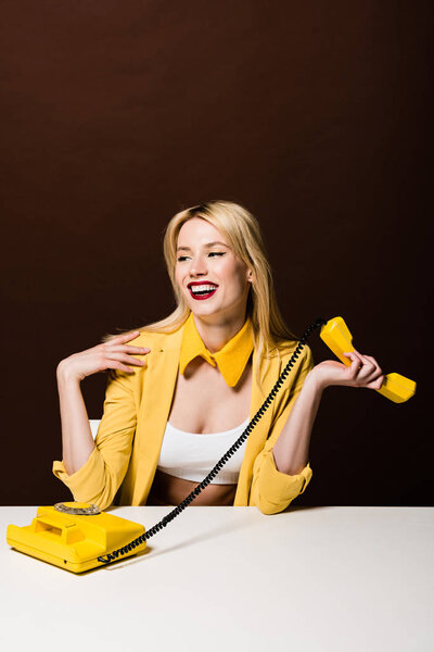 beautiful smiling blonde girl holding yellow handset and looking away on brown