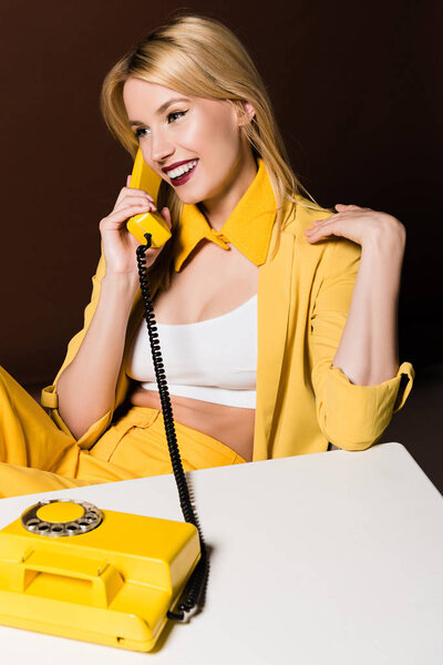 beautiful smiling stylish blonde girl talking by yellow vintage phone on brown