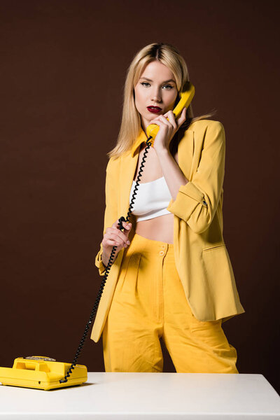 beautiful blonde girl talking by yellow vintage phone and looking at camera on brown