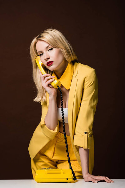 beautiful young blonde woman talking by vintage phone and looking at camera on brown