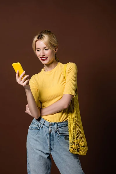Piękna Blondynka Uśmiechający Się Ciąg Worek Żółty Smartfona Brązowym Tle — Zdjęcie stockowe