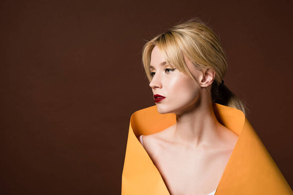 beautiful blonde woman wrapped in orange paper looking away isolated on brown 