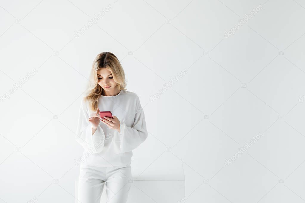 portrait of blond woman in white clothing using smartphone isolated on grey