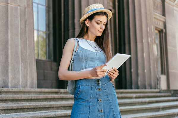 attractive smiling girl in hat using digital tablet while standing on stairs