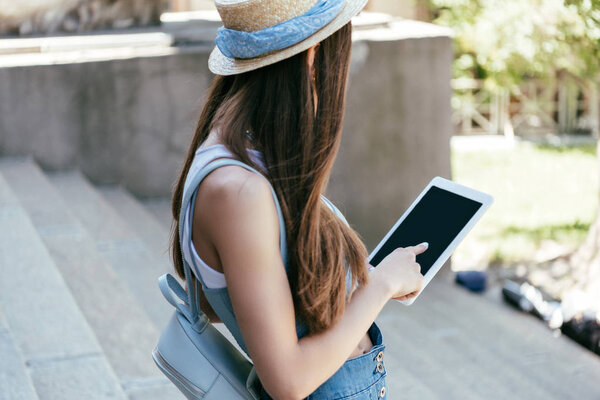 side view of young woman in hat using digital tablet with blank screen on stairs 