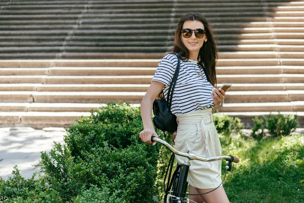 Beautiful Young Woman Holding Smartphone Smiling Camera While Standing Bicycle — Free Stock Photo