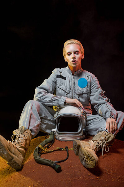 beautiful female cosmonaut in spacesuit holding helmet and sitting on planet