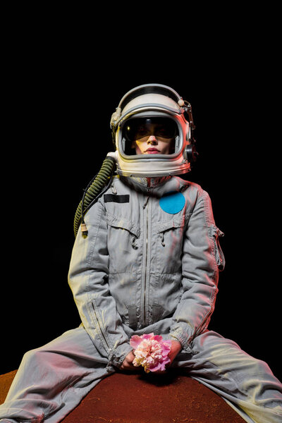 female astronaut in spacesuit and helmet sitting on planet with flower in cosmos