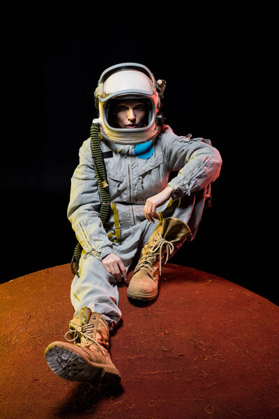 young cosmonaut in spacesuit with helmet sitting on planet in space