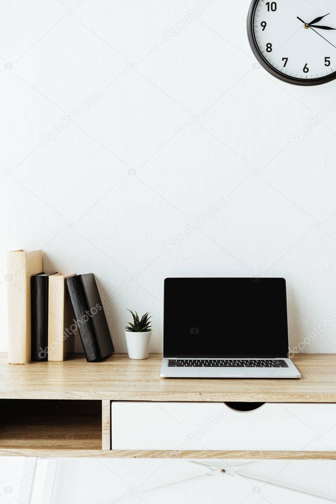 laptop with blank screen and books on work desk in front of white wall