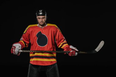 professional ice hockey player holding hockey stick and looking at camera isolated on black clipart