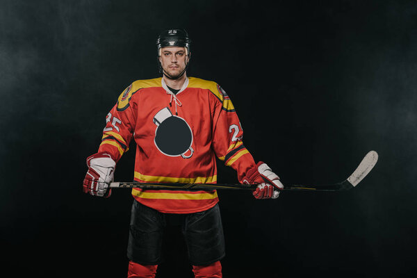 professional ice hockey player holding hockey stick and looking at camera isolated on black 