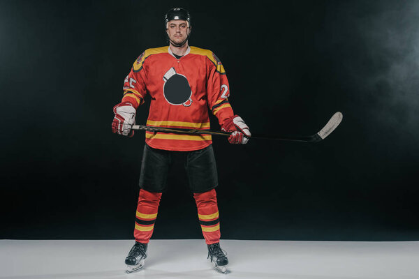 full length view of professional sportsman in skates holding hockey stick and looking at camera on black